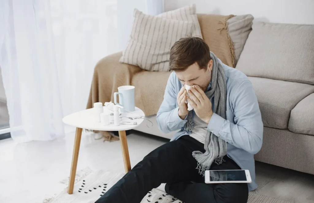 Man sitting on the floor sneezing with tissue near sofa, showing allergy or illness symptoms caused by poor indoor air quality.