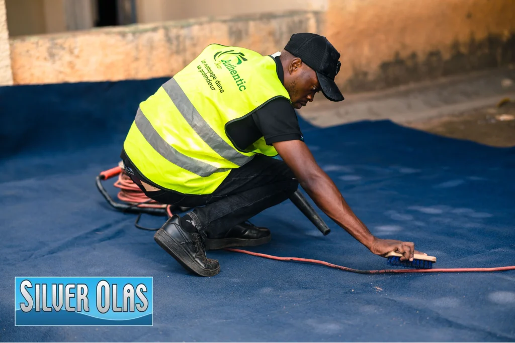 Worker cleaning blue flooring with brush while wearing safety vest during professional floor maintenance.