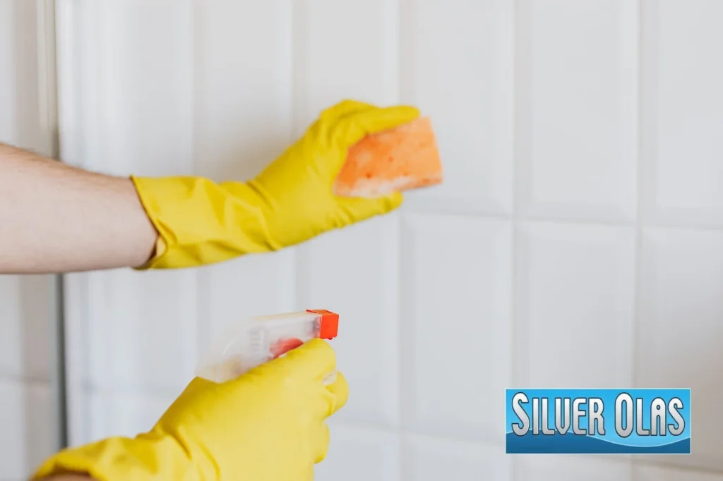 Hands in yellow rubber gloves using a spray bottle and sponge for professional tile cleaning in Carlsbad on white ceramic wall tiles.